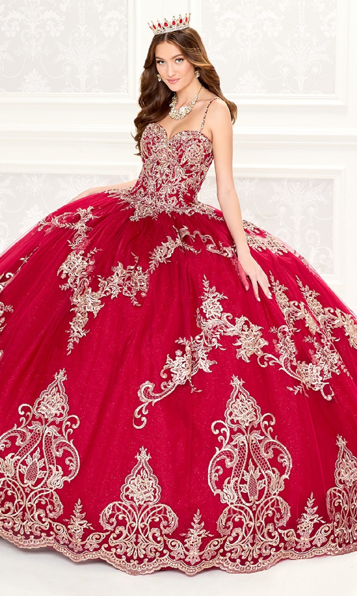 A young woman in the Quinceanera Dress PR30086 By Princesa, featuring gold embroidery and a jeweled crown, stands against an elegant white background.